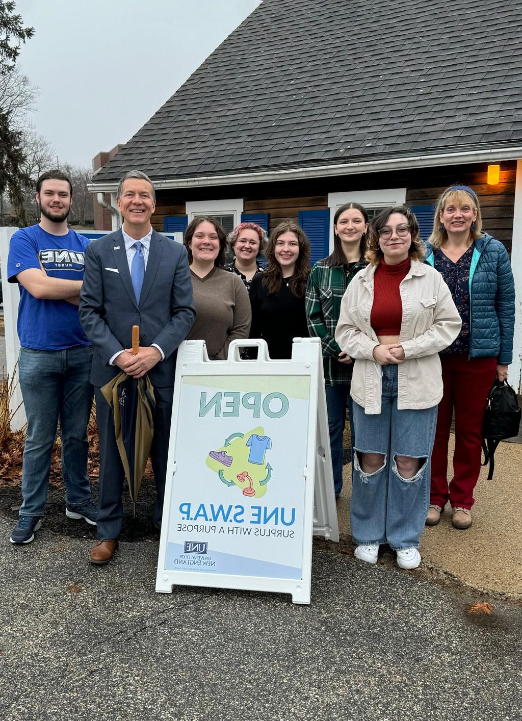 President Herbert stands with U N E students behind an "Open" sign for the Swap 商店