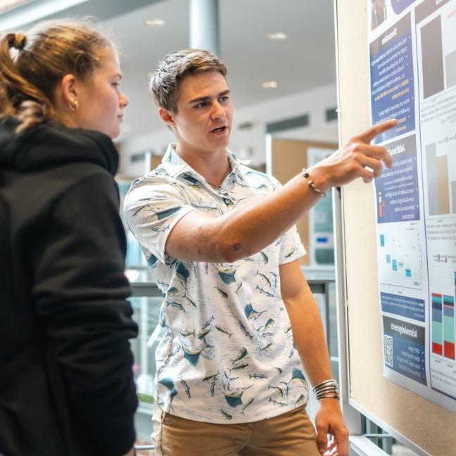 A student points to their presentation poster as they talk to a fellow student