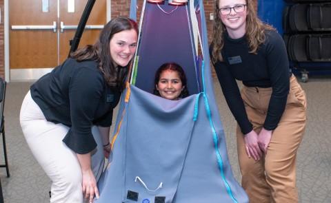 A child sits inside a suspended tent and two students pose around her